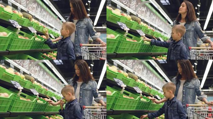 可爱的小男孩正在杂货店的水果和蔬菜部门选择苹果，他迷人的母亲站在他旁边和她的儿子说话。