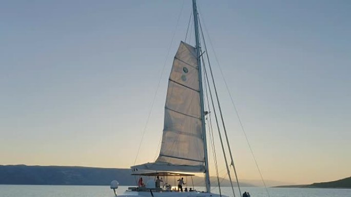 一艘帆船双体船的空中拍摄，帆船在平静的海面上航行，太阳升起在山后。