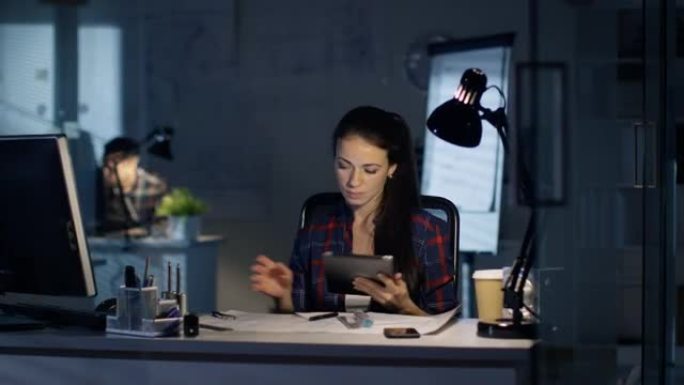 在深夜办公室，女设计工程师负责草稿，她咨询平板电脑。她的桌子被灯照亮了。同事在后台工作。