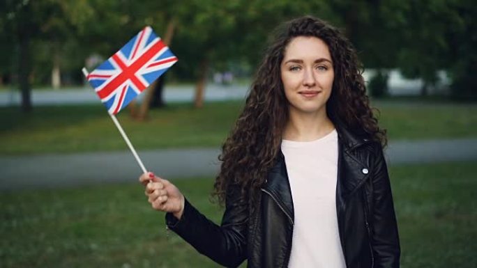 快乐的英国学生挥舞着英国国旗的慢动作肖像，看着相机，微笑着站在公园的户外。人和国家概念。