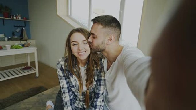 年轻微笑和美丽的夫妇的POV在智能手机相机上自拍，早上坐在床上亲吻