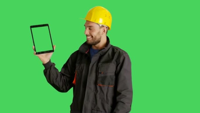 一个英俊的工人戴着安全帽，用一只手拿着平板电脑，用另一只手滑动触摸手势。平板电脑和背景是绿色屏幕。