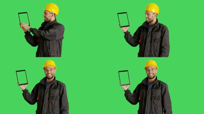 一个英俊的工人戴着安全帽，用一只手拿着平板电脑，用另一只手滑动触摸手势。平板电脑和背景是绿色屏幕。