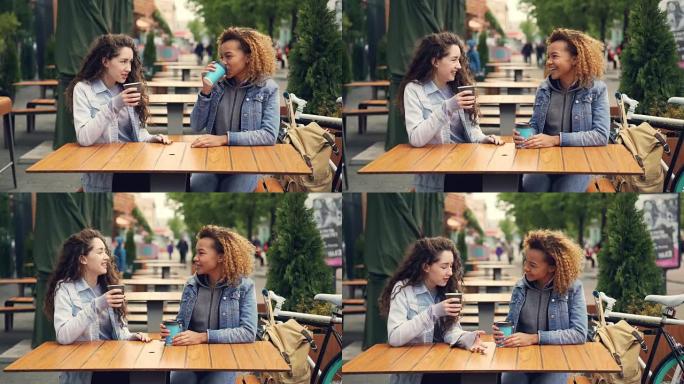 两个年轻女人坐在公园户外咖啡馆的餐桌旁喝咖啡和聊天，年轻女人开心又笑。谈话和饮料概念。