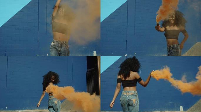 美丽的年轻女子手持五颜六色的烟雾弹对着蓝墙在外面跳舞