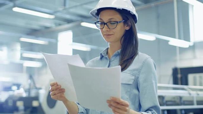 戴着安全帽的女工程师站在大工厂中间时会带着蓝图工作。