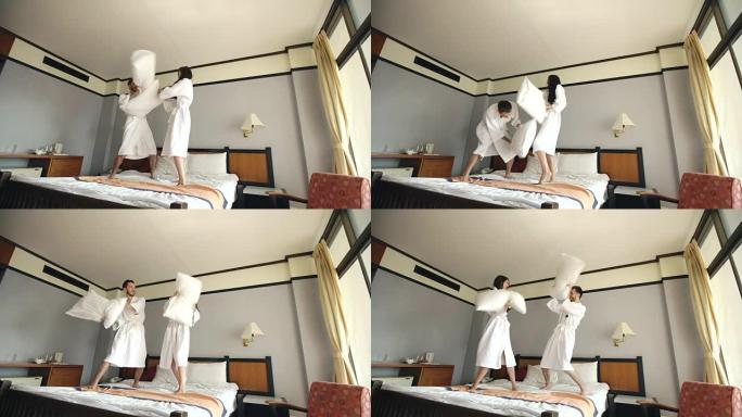 穿着浴袍的年轻幸福夫妇在蜜月假期期间在酒店的床上打架枕头，玩得开心