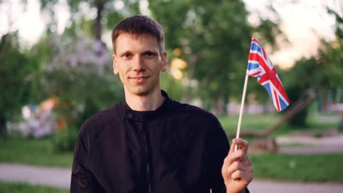 微笑的年轻人骄傲的公民手持英国国旗站在街道上的慢动作肖像，背景是尼斯公园，看着相机。