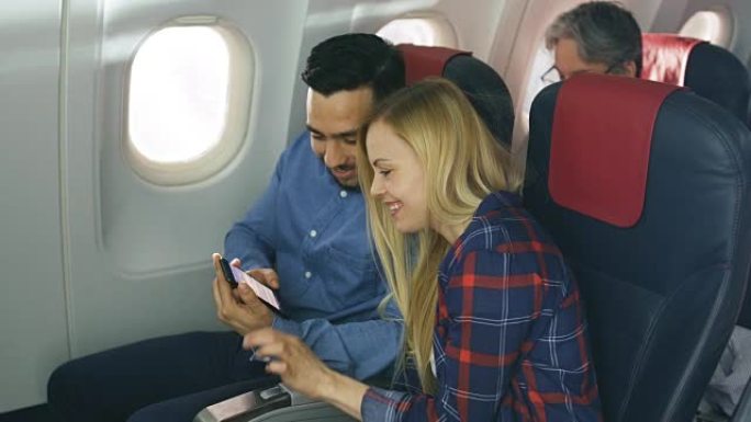 在商用飞机上，漂亮的年轻金发女郎与英俊的西班牙裔男性在智能手机上观看社交媒体并大笑。高级乘客看书。