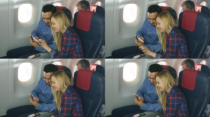 在商用飞机上，漂亮的年轻金发女郎与英俊的西班牙裔男性在智能手机上观看社交媒体并大笑。高级乘客看书。