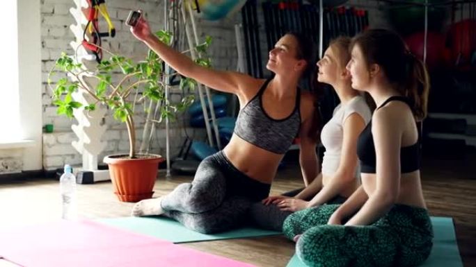 瑜伽学生坐在瑜伽馆明亮的垫子上时，正在用智能手机自拍。女人摆姿势，微笑着大笑。健康乐趣概念。