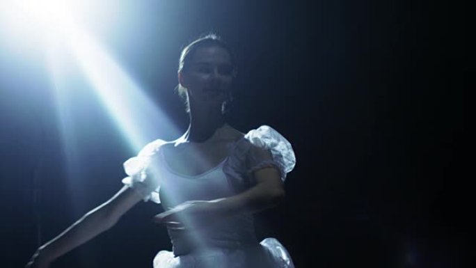 一位美丽的年轻芭蕾舞演员在聚光灯下优雅地跳舞，周围是黑暗。她穿着白色短裙，在灯光下闪闪发光。慢动作。