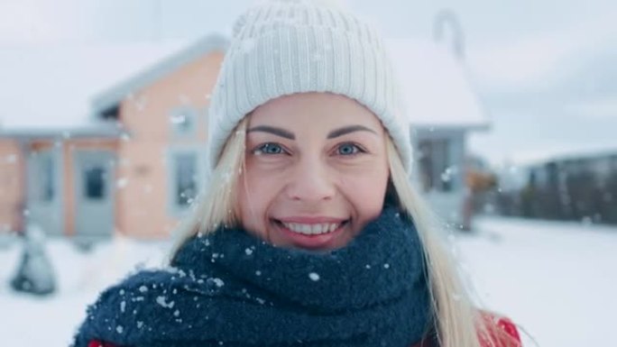 一位年轻的金发女子穿着红色外套，戴着围巾，站在飘落的雪中，对着镜头微笑。在后院的田园诗般的房子里，可