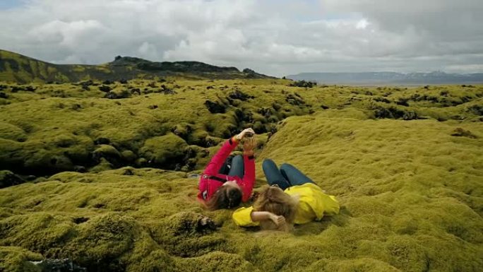 两名妇女躺在冰岛软熔岩场上的鸟瞰图。在智能手机上自拍的游客