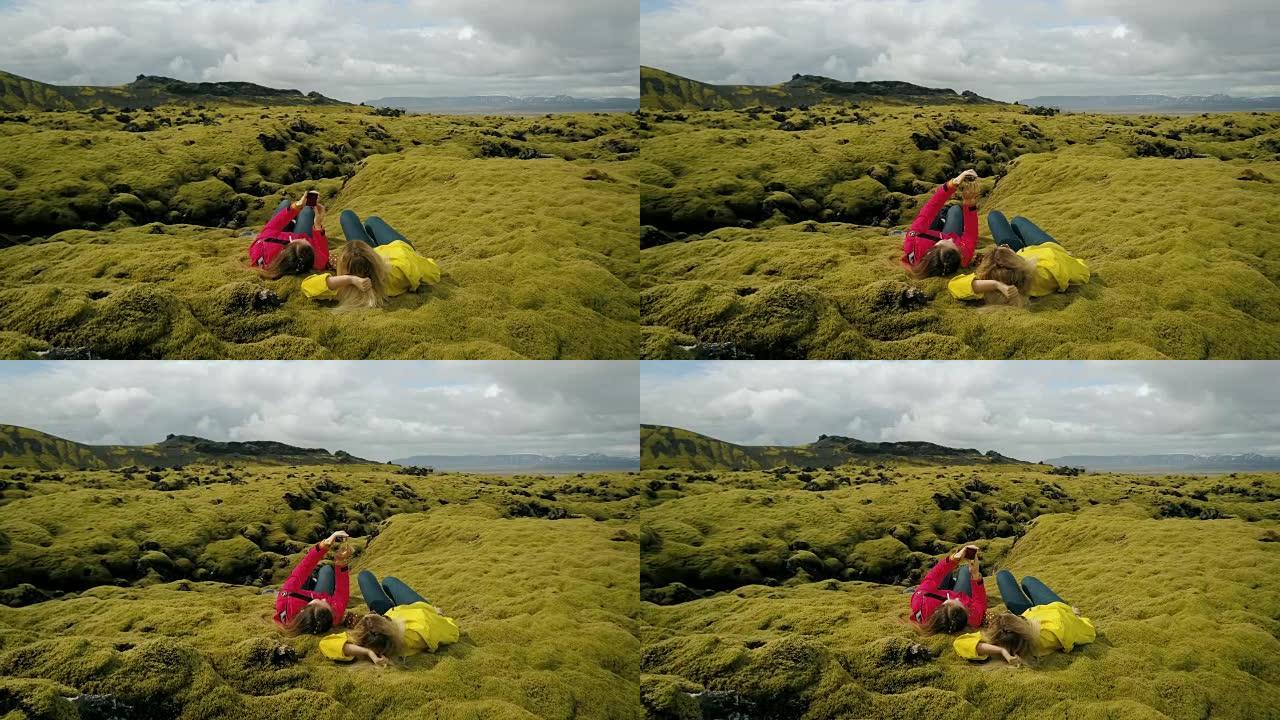 两名妇女躺在冰岛软熔岩场上的鸟瞰图。在智能手机上自拍的游客