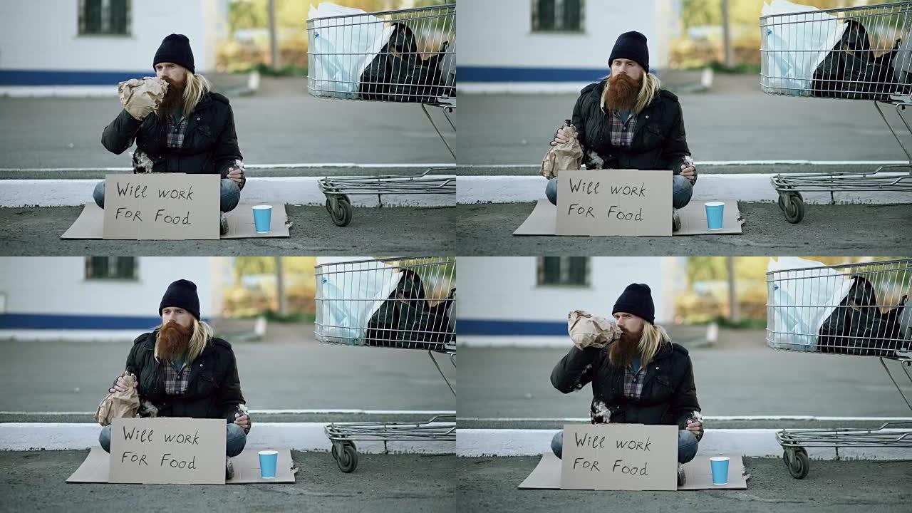 大胡子的年轻无家可归的人，拿着纸板坐在购物车附近，在寒冷的日子里喝酒