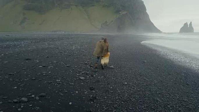 在冰岛巨魔脚趾附近的黑色火山海滩上奔跑的年轻时髦夫妇的后鸟瞰图