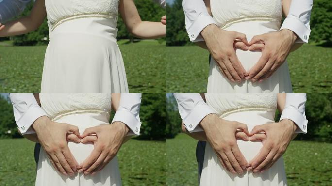 特写: 无法识别的夫妻用手在怀孕的腹部上使爱心