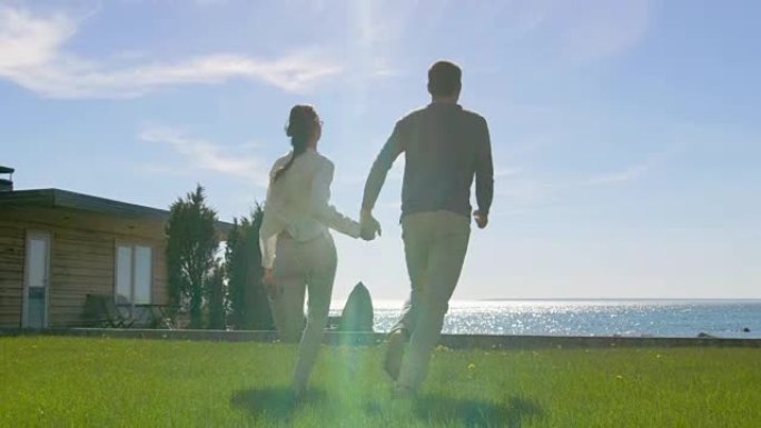 在美丽的夏日，幸福的夫妻手牵手，赤脚在草地上奔跑。在他们面前阳光明媚的蓝天和海滨景色。