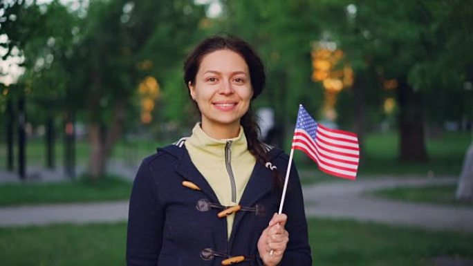 骄傲的美国公民微笑的女人挥舞着美国国旗，看着相机微笑的慢动作肖像。可以看到公园里的绿树，草坪，小路。