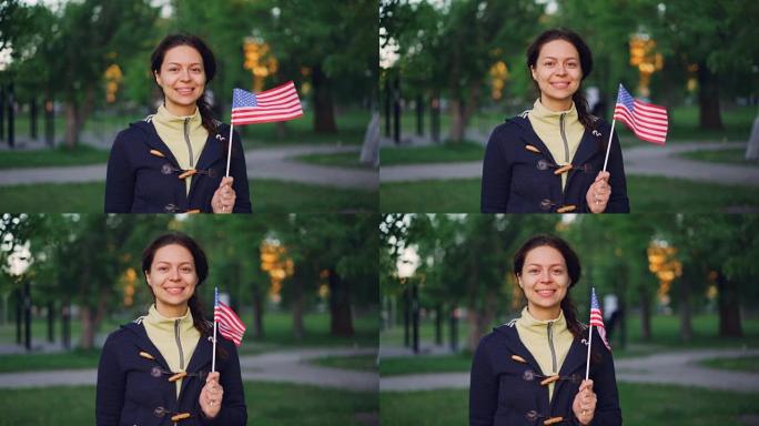 骄傲的美国公民微笑的女人挥舞着美国国旗，看着相机微笑的慢动作肖像。可以看到公园里的绿树，草坪，小路。