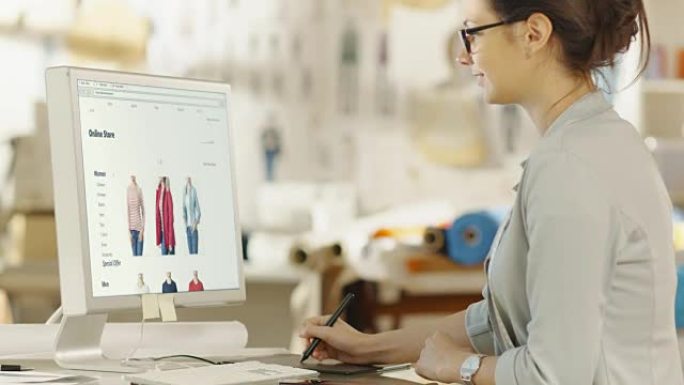 女性时装设计师在她的个人电脑上使用数字绘图板，她为在线零售店设计新网站。背景阳光明媚的工作室里有面料