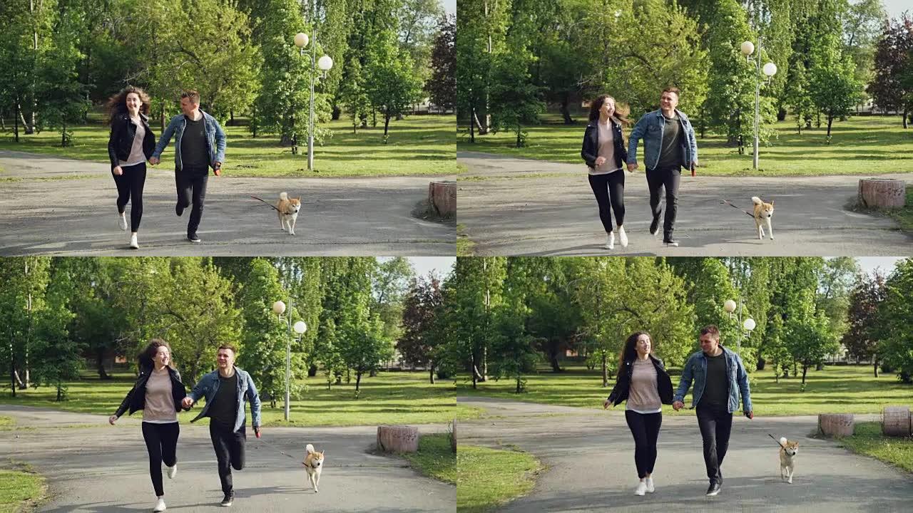 年轻的已婚夫妇在公园里跑步的慢动作，小狗享受自由和活动，周围有美丽的绿树和草丛。