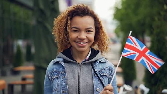在大风天，年轻漂亮的非洲裔美国女士拿着英国国旗微笑着看着相机的慢动作肖像。快乐的游客和有吸引力的人的