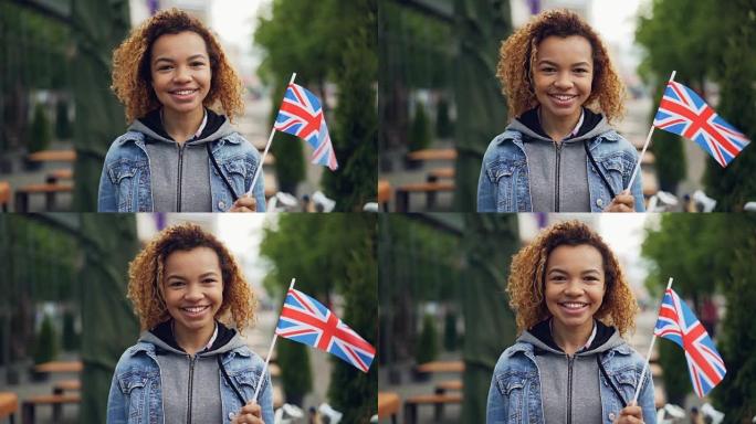在大风天，年轻漂亮的非洲裔美国女士拿着英国国旗微笑着看着相机的慢动作肖像。快乐的游客和有吸引力的人的
