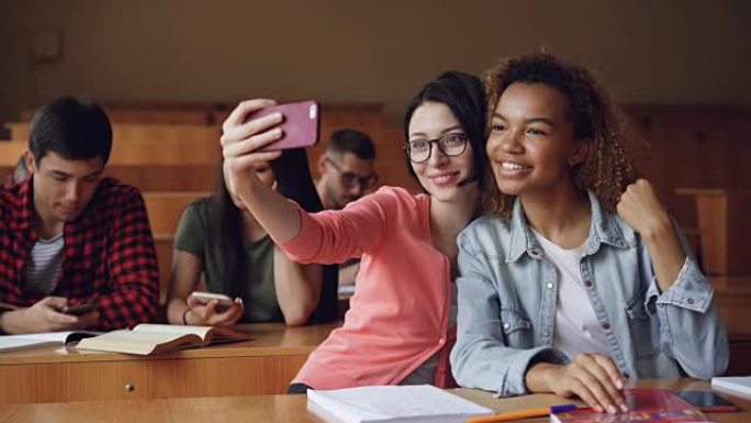 开朗的女生坐在大学里的桌子上，用智能手机自拍，女人摆姿势做手势和拥抱。朋友和技术概念。