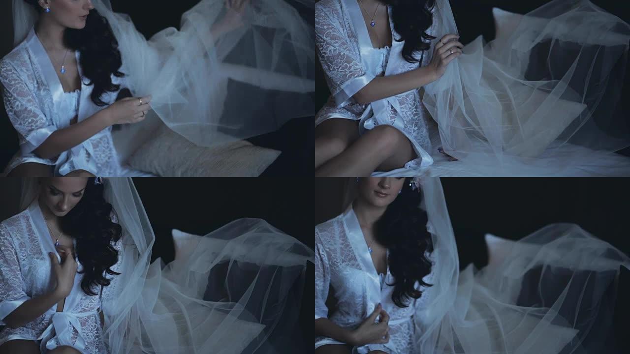 年轻的黑发新娘穿着优雅的白色内衣，peignoir坐在床上。美丽的女人修理她的面纱