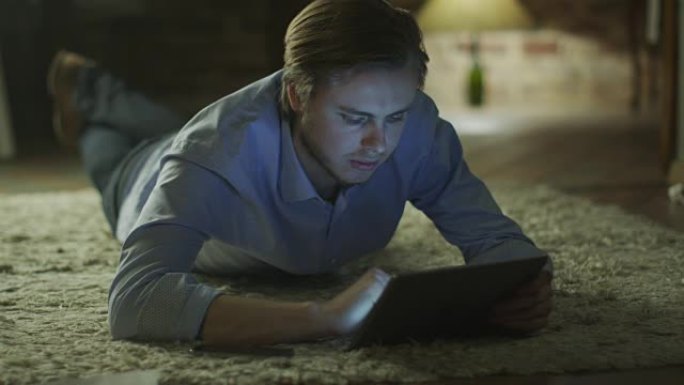 年轻人躺在地板上，晚上在家使用平板电脑娱乐。休闲生活方式。