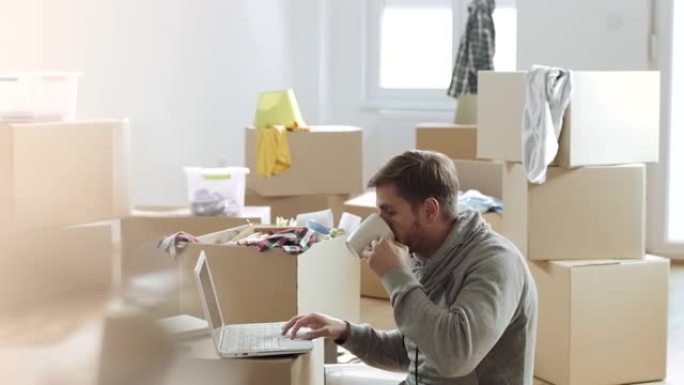 在装满未包装盒子的新公寓里工作的人在笔记本电脑上喝咖啡