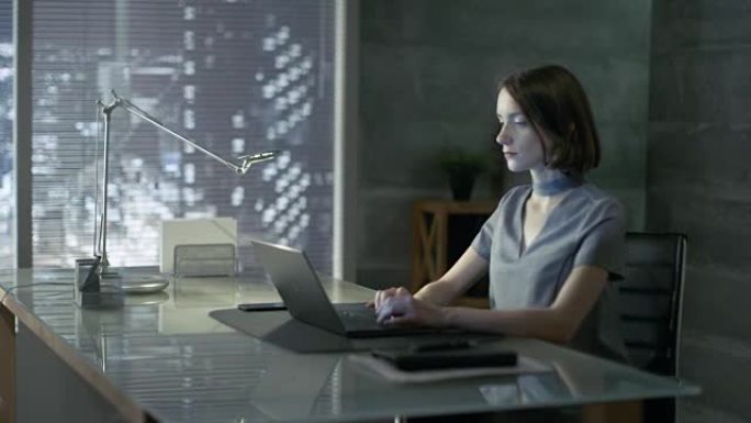 成功的年轻女子坐在办公桌前的私人办公室里，在笔记本电脑上工作。她的办公室以极简主义风格完成，很暗，窗