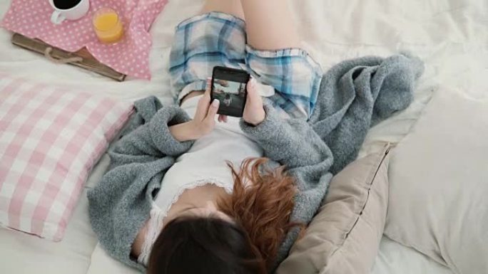 年轻漂亮的女人躺在床上，拿着手机。女孩在智能手机上观看旅行中的照片