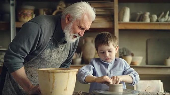 有爱心的祖父正在看着他的孙子在投掷轮上模制粘土，制作低矮的陶瓷花瓶，然后在大碗中洗手。陶器和家庭概念