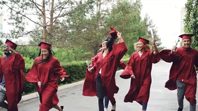 情绪激动的毕业生的慢动作快乐的女孩和男孩带着文凭奔跑并大笑，漂亮女孩正在脱下迫击炮弹并挥舞着它。可见