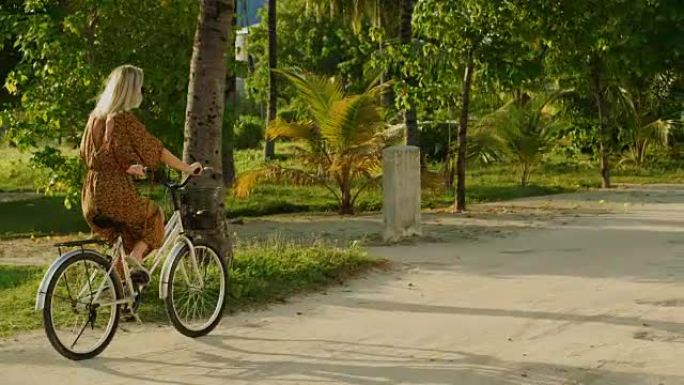 穿着连衣裙的美女骑着自行车在路上穿过正宗的村庄。南亚自然，风景秀丽的森林在路边生长。