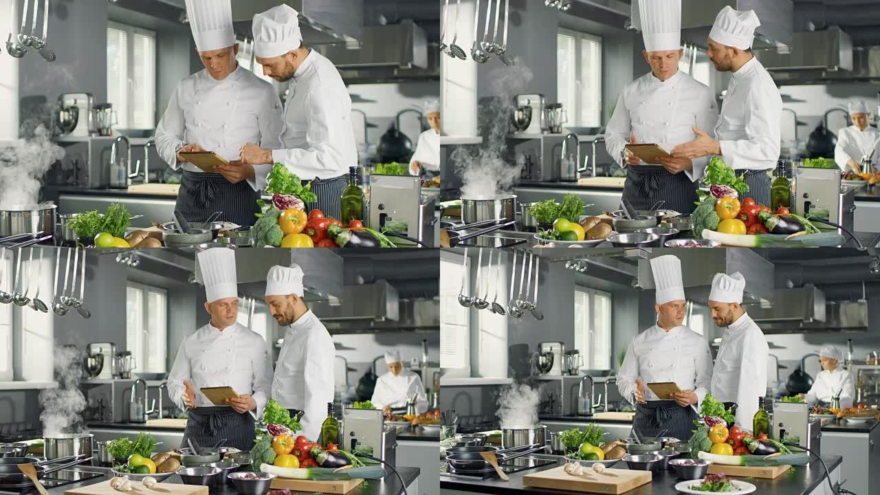 两位著名厨师在使用平板电脑时讨论他们的视频博客。他们在大型餐厅不锈钢专业厨房工作。