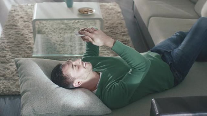 男子躺在家里的沙发上，穿着牛仔裤和绿色外套，使用智能手机。