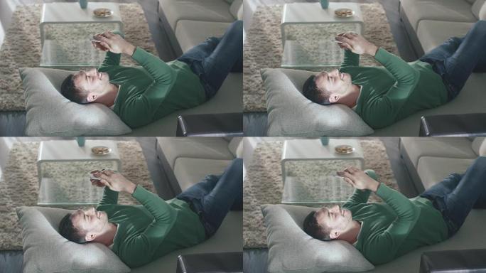 男子躺在家里的沙发上，穿着牛仔裤和绿色外套，使用智能手机。