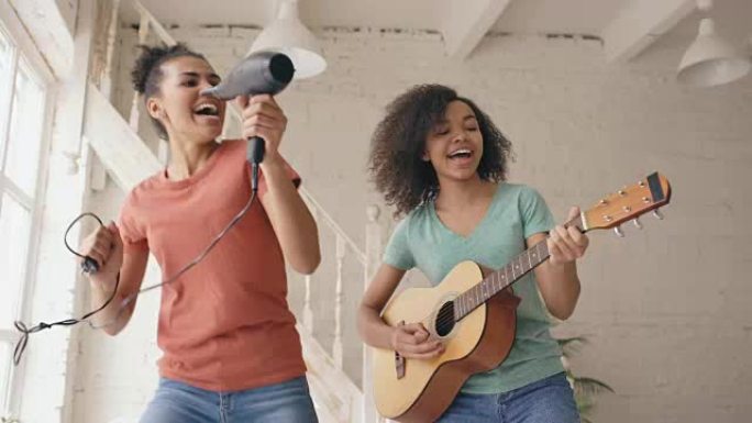 混合种族年轻有趣的女孩在床上用吹风机跳舞唱歌和弹吉他。姐妹们在家里的卧室里玩得开心