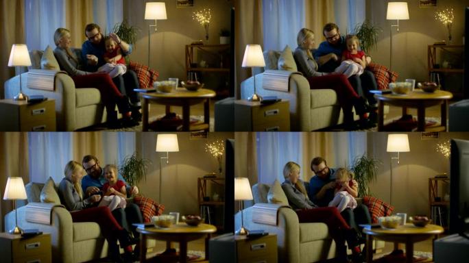 父亲、母亲和小女孩看电视的长镜头。父亲向他的小女孩解释了一些事情。他们坐在舒适的客厅的沙发上。今天晚
