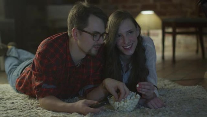年轻快乐的微笑夫妇躺在地板上，看电视，吃爆米花。