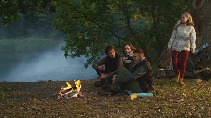一群朋友坐在篝火旁湖边的森林里，喝着热饮，而另一个女孩则从帐篷里走出来。
