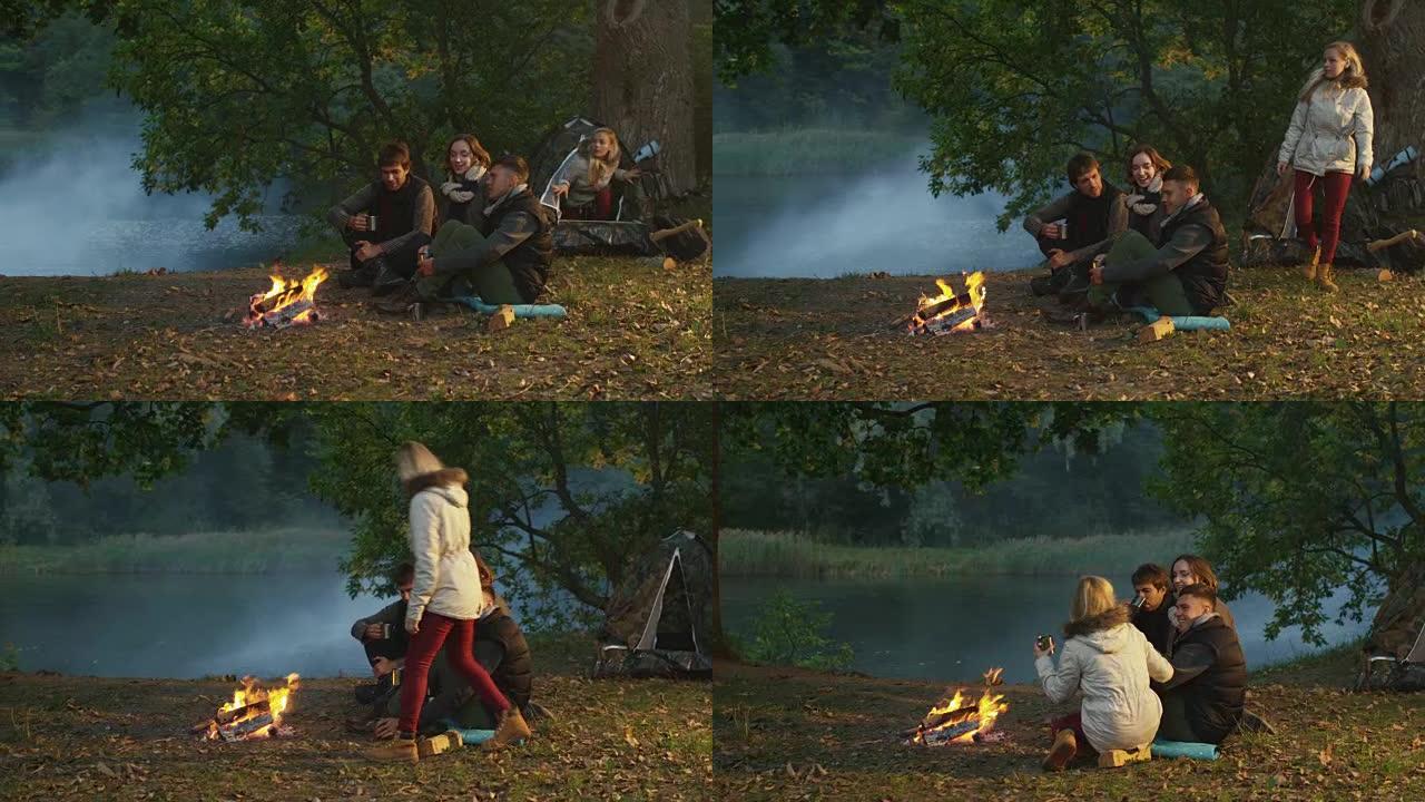 一群朋友坐在篝火旁湖边的森林里，喝着热饮，而另一个女孩则从帐篷里走出来。