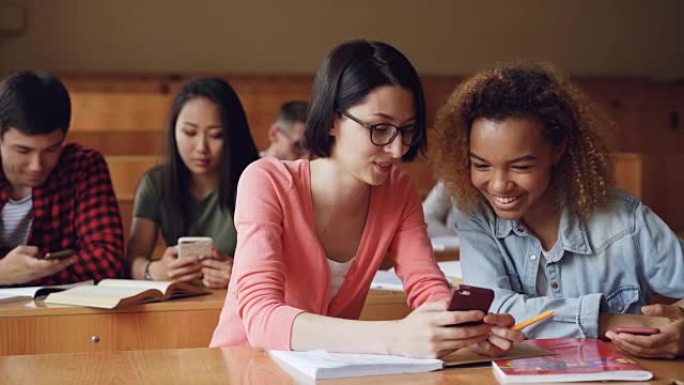 漂亮女孩的学生正在使用智能手机，看屏幕，坐在大学的课桌上聊天和大笑。社交媒体、互联网、千禧一代和教育