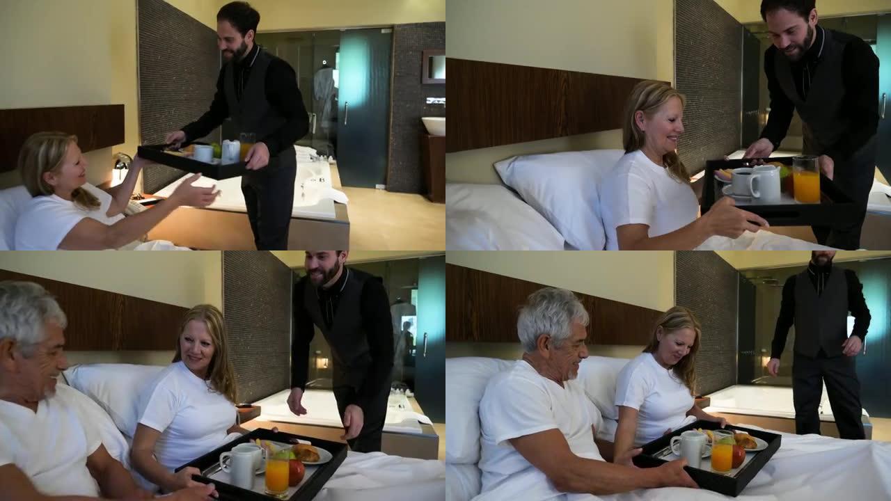 友好的服务员在酒店微笑着为一对躺在床上的老年夫妇带来客房服务早餐