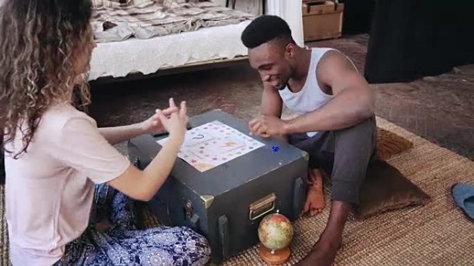 多种族夫妇穿着睡衣坐在地板上，一起玩棋盘游戏。男人扔骰子拿卡