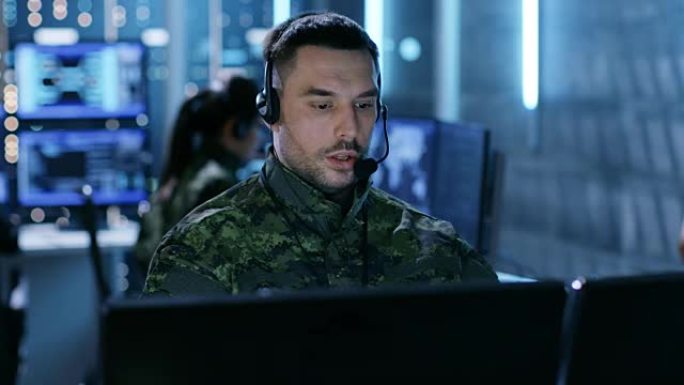军事技术支持专业人员使用耳机进行指导。他在一间监控室里，还有其他军官和许多后台工作显示器。